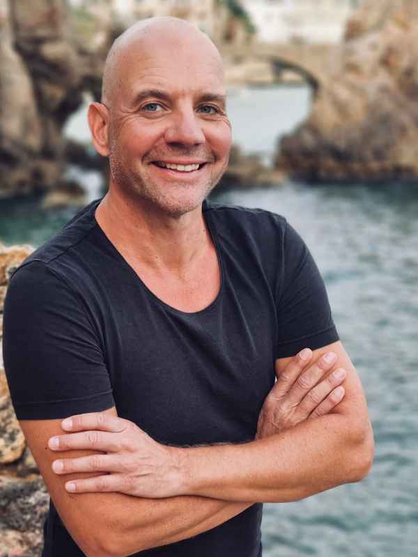 Male Escort Sascha from haut-nah.org, standing by the sea in front of a rock, smiling, with his arms crossed