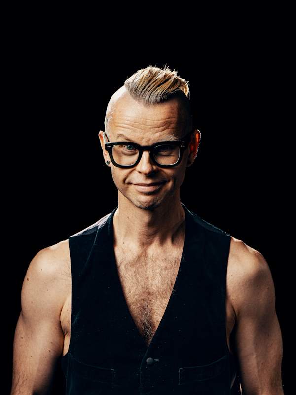Callboy Mann Tom von haut-nah.org mit schwarzer Weste und freien Oberkoerper und Brille, lächelt