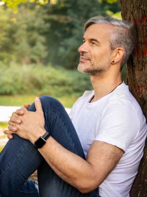 Callboy Roman von haut-nah.org im Sitzen von der Seite am Baum gelehnt, lächelnd