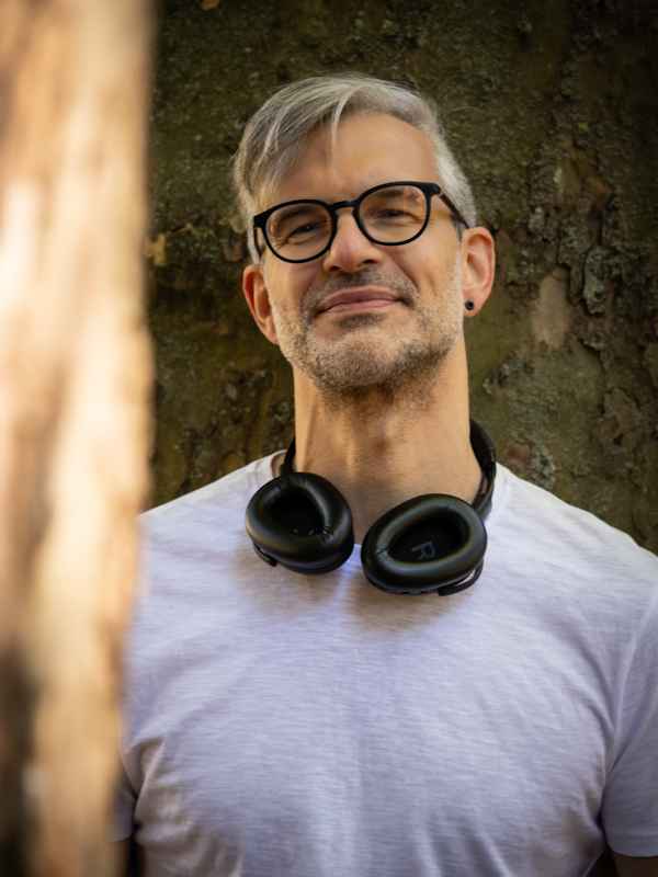 Callboy Roman von haut-nah.org mit Kopfhörer um Hals am Baum gelehnt, lächelnd