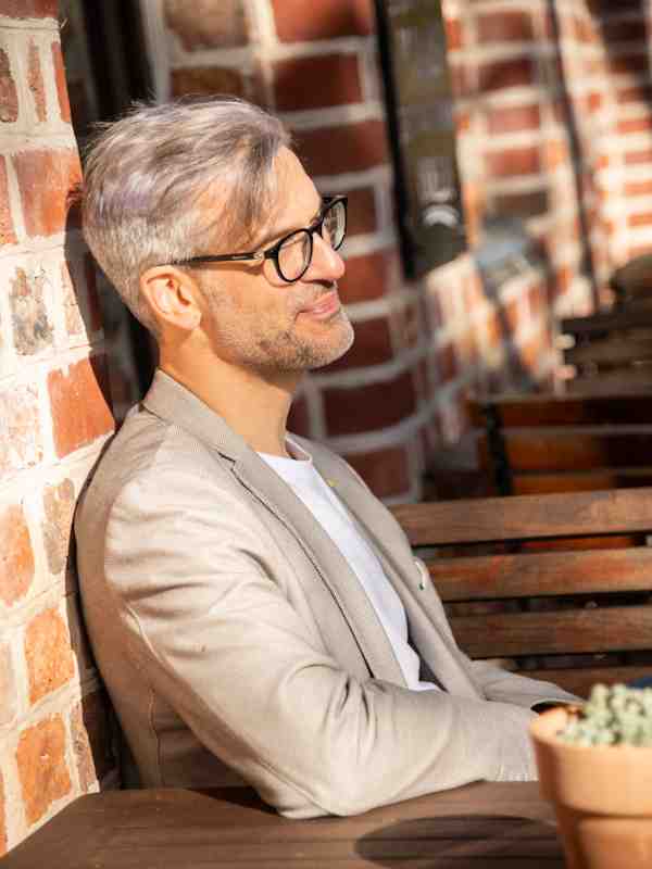 Escort Mann Roman von haut-nah.org seitlich mit beigen Sakko an Ziegewand gelehnt