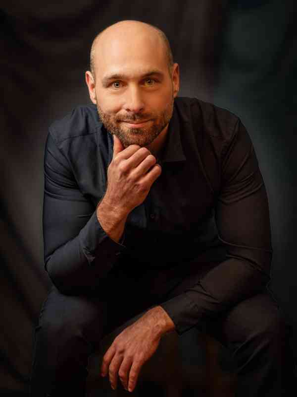 Callboy William von haut-nah.org im Sitzen, leicht nach vorne gebeugt im schwarzen Hemd