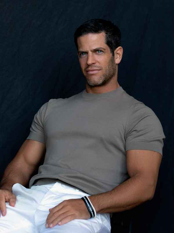 Male Escort Chris from haut-nah.org sitting in white pants and brown shirt, smiling slightly