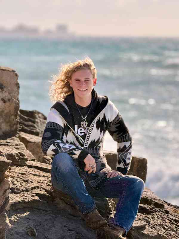 Callboy Xavier von haut-nah.org in Skandinavierpulli, sitzt auf Felsen, lächelt