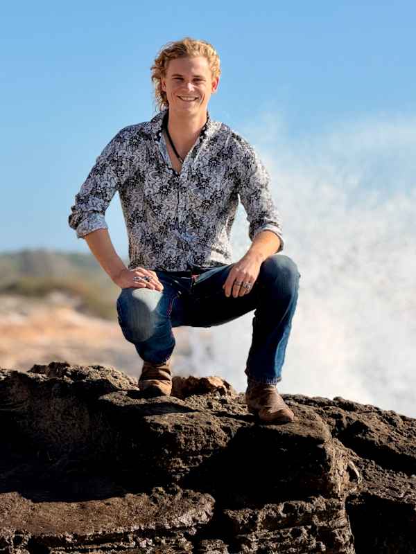 Escort Mann Xavier von haut-nah.org in Jeans und bunten Hemd knieend auf Felsen vor dem Meer