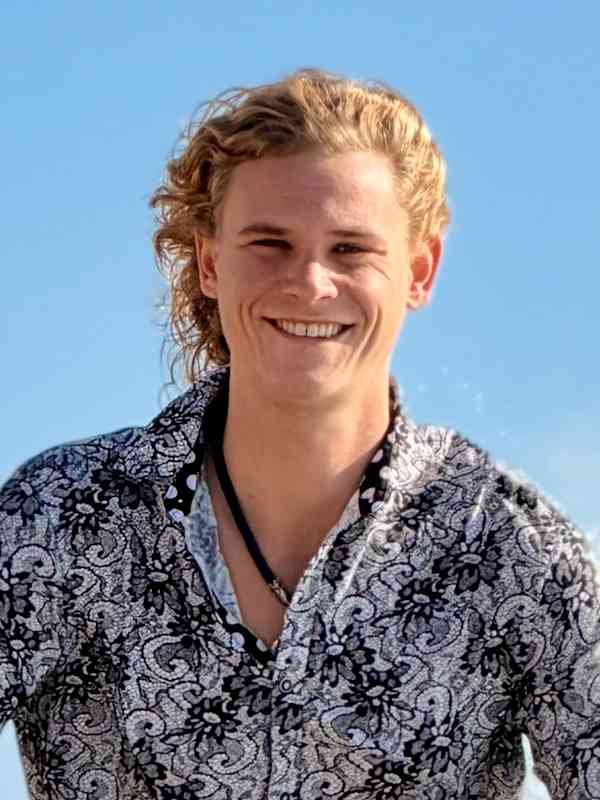 Escort Mann Xavier von haut-nah.org in Jeans und bunten Hemd knieend auf Felsen vor dem Meer, Nahaufnahme