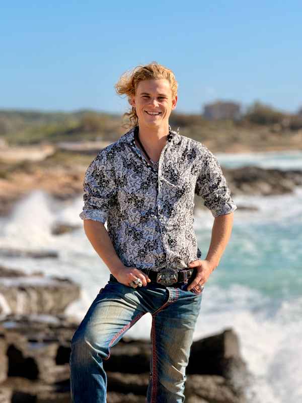 Escort Mann Xavier von haut-nah.org in Jeans und bunten Hemd stehend, Knie nach vorne auf Felsen vor dem Meer