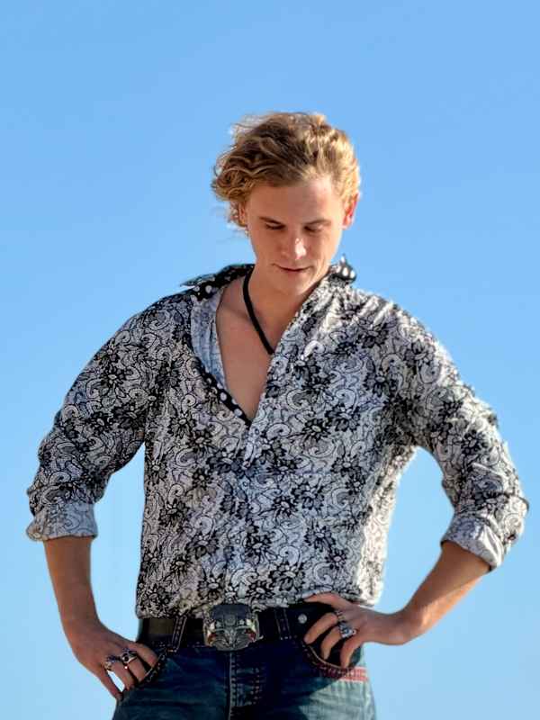 Escort Mann Xavier von haut-nah.org in Jeans und bunten Hemd steht auf Felsen, Hände in den Hüften, Kopf gesenkt