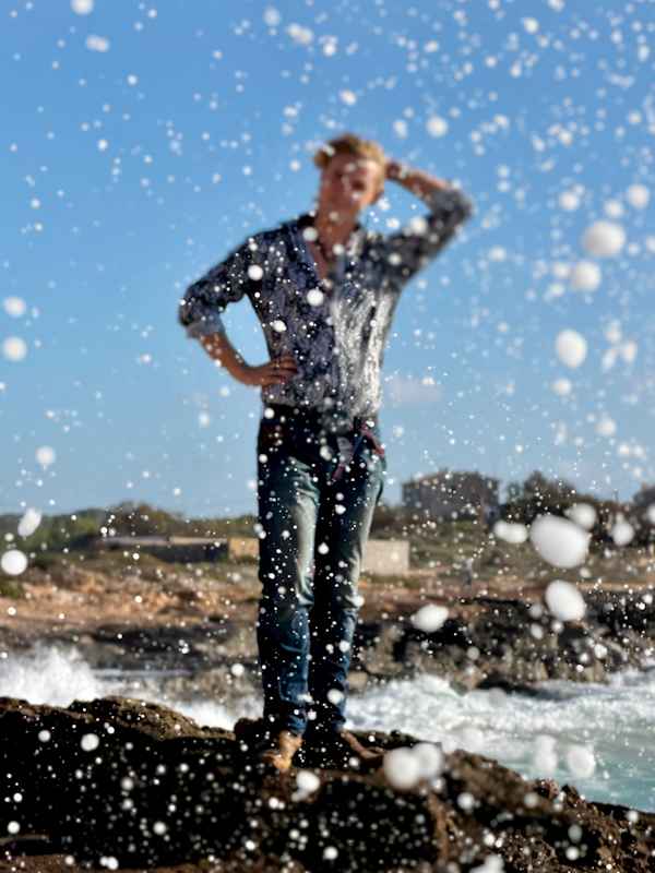 Escort Mann Xavier von haut-nah.org in Jeans und bunten Hemd steht auf Felsen, verschwommen, mit Wasser auf der Kameralinse