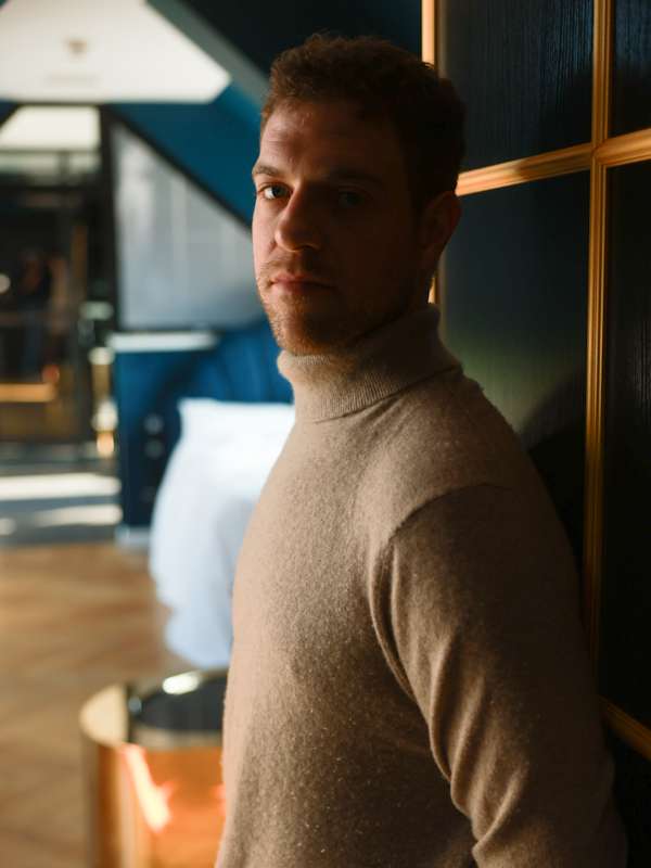 Escort Mann Simon von haut-nah.org an Wand gelehnt mit Rollkragenpulli